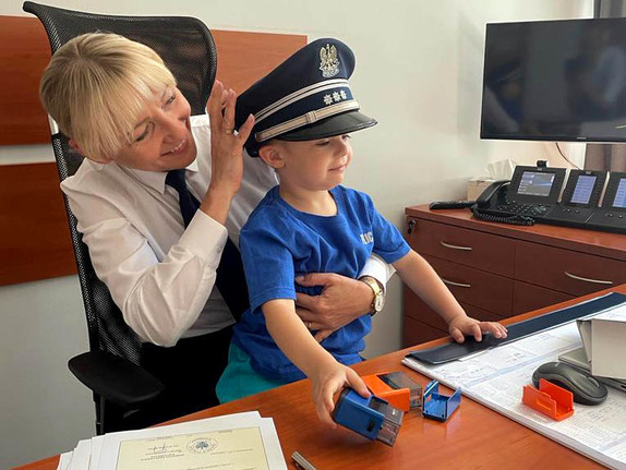 Dziecko w czapce policyjnej przy biurku w gabinecie razem z zastępcą komendanta insp. dr Gabrielą Sochą